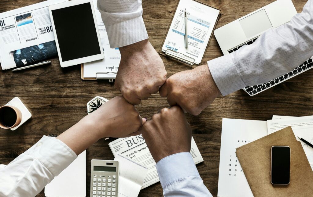 Four different office workers are bumping their fists together over a meeting desk. On the desk are phones, calculators, tables, and various documents. They are celebrating a successful meeting.